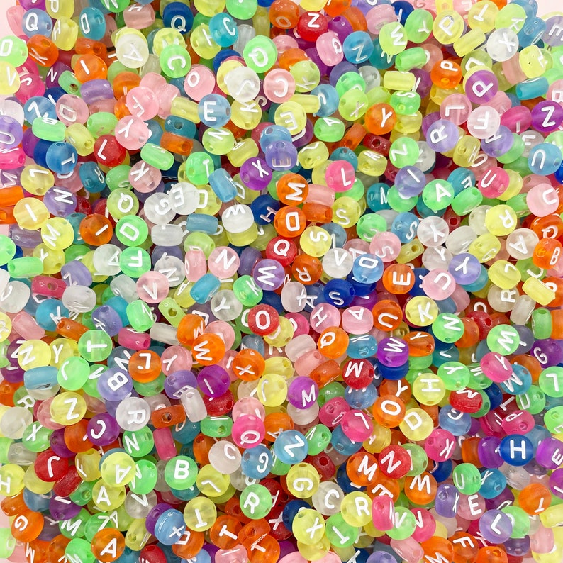 May include: A close-up of a large pile of colourful alphabet beads. The beads are translucent and have white letters on them. The beads are a mix of colours, including pink, blue, green, yellow, orange, and purple.