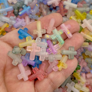 Puede incluir: Una colección de pequeños abalorios en forma de cruz, multicolores, en tonos pastel, incluyendo rosa, azul, amarillo y verde. Algunas cruces tienen motas doradas. Los abalorios son de un material translúcido.