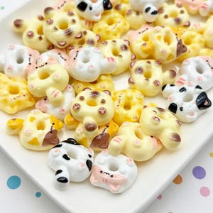 May include: A collection of small, cat-shaped donuts in various colors, including yellow, white, and brown, arranged on a white rectangular plate. The donuts have cute facial features and paw prints.