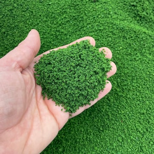 May include: A close-up of a hand holding a pile of green artificial turf. The turf is made of small, green granules.