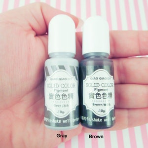 Pu&ograve; includere: Due piccoli flaconi di pigmento colorato solido grigio e marrone. Ogni flacone &egrave; etichettato "QIAO QIAO DIY SOLID COLOR Pigment 實色色精" e include il nome del colore in inglese e cinese. Il flacone grigio dice "Gray/灰色 -10g-" e il flacone marrone dice "Brown/棕色 -10g-".