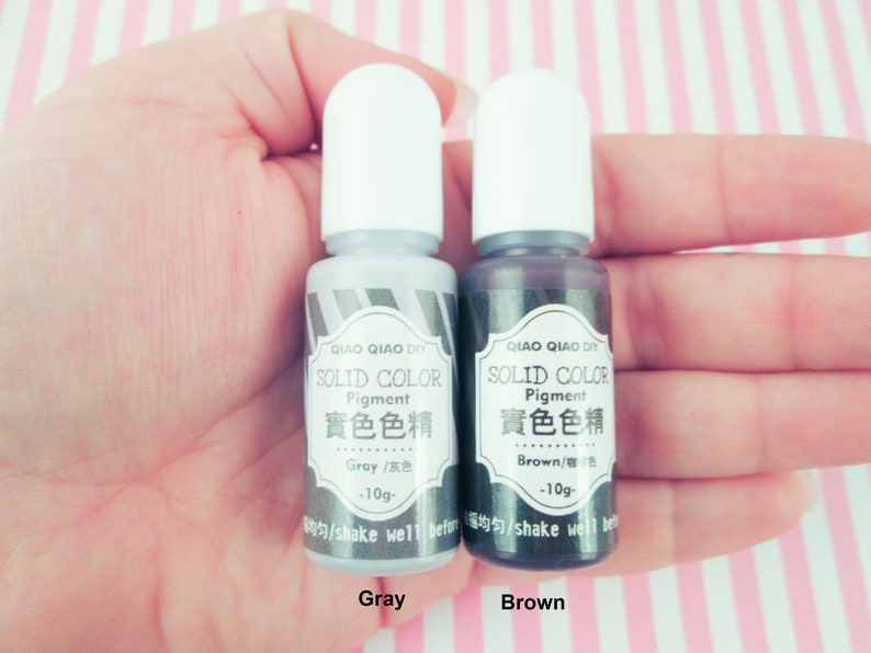 Pu&ograve; includere: Due piccoli flaconi di pigmento colorato solido grigio e marrone. Ogni flacone &egrave; etichettato "QIAO QIAO DIY SOLID COLOR Pigment 實色色精" e include il nome del colore in inglese e cinese. Il flacone grigio dice "Gray/灰色 -10g-" e il flacone marrone dice "Brown/棕色 -10g-".