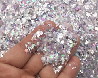 Puede incluir: Primer plano de copos de purpurina iridiscente en blanco, plata y lavanda. Los copos, de forma irregular, se sostienen en una mano, revelando su textura y capacidad para reflejar la luz. Efecto brillante.