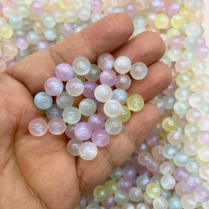 May include: A hand holding a variety of iridescent pastel-colored glass beads. The beads are round and have a shimmery finish.