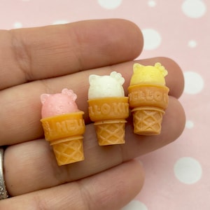 May include: Three miniature ice cream cones with different colored ice cream. The ice cream is shaped like a cartoon animal head. The cones are brown and have a waffle pattern. The ice cream flavors are pink, white, and yellow.