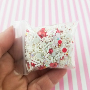 May include: A clear plastic bag filled with white and red sprinkles, including snowflake shapes, small balls, and white sticks.