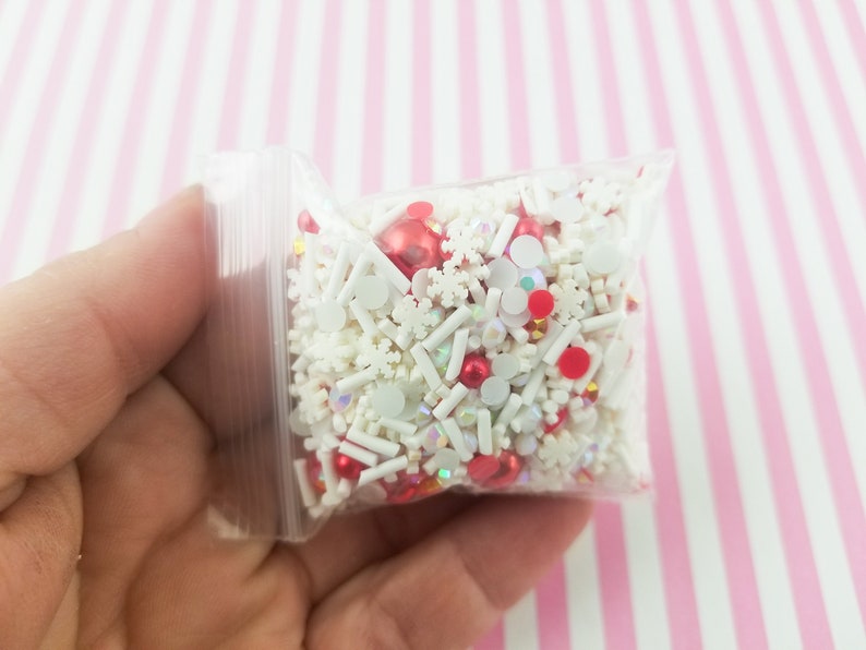 May include: A clear plastic bag filled with white and red sprinkles, including snowflake shapes, small balls, and white sticks.