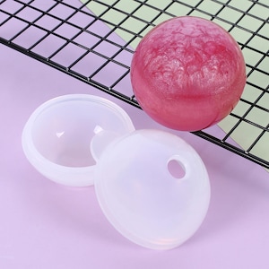 May include: A white silicone ice ball mould with a lid, next to a large, textured, pink ice sphere. The mould is open, revealing the interior. The items are on a purple surface, with a black wire rack in the background.