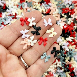 May include: Assortment of small plastic bows in various colors, including red, white, black, and pink. The bows are scattered on a surface and a hand is holding a few of them.