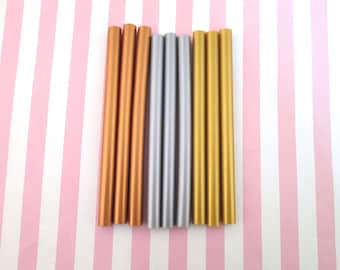 May include: Eight sticks of sealing wax in three colors: gold, copper, and silver. The sticks are arranged in a row on a pink and white striped background.