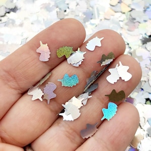 A hand holding a collection of small, colorful unicorn-shaped glitter pieces against a background of glitter.