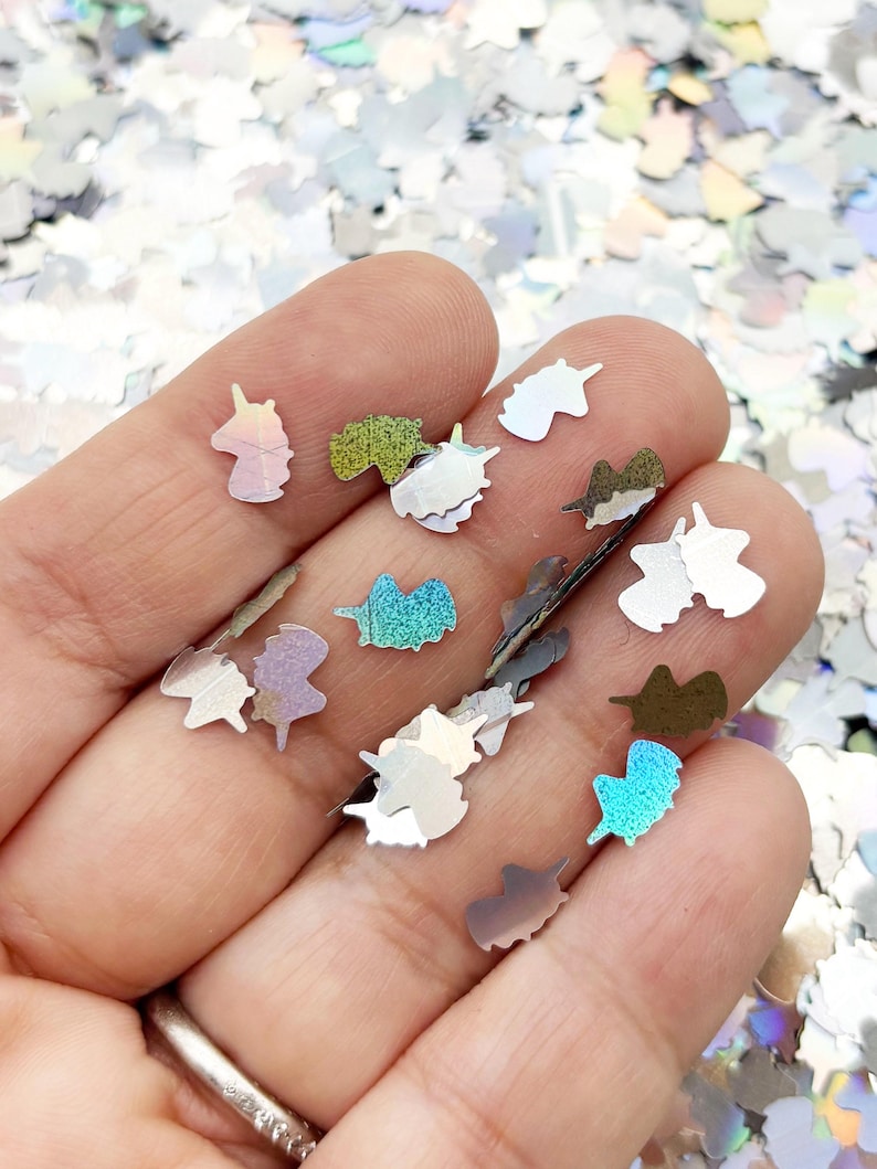 A hand holding a collection of small, colorful unicorn-shaped glitter pieces against a background of glitter.