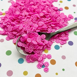 May include: A close-up of a spoon filled with pink and white plastic citrus fruit slices. The spoon is on a white background with colourful polka dots.