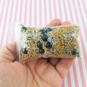 May include: A clear plastic bag filled with gold and black sprinkles. The sprinkles are a mix of small gold rods, clear square sprinkles, and black round sprinkles.