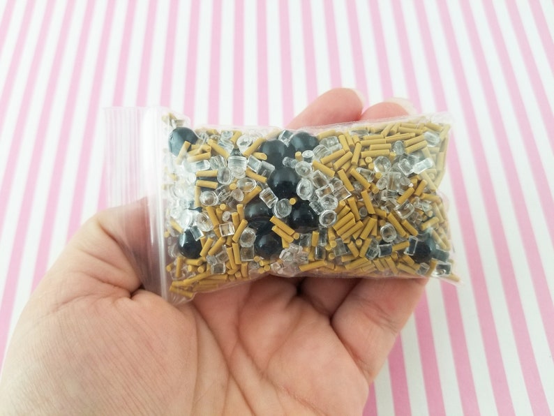 May include: A clear plastic bag filled with gold and black sprinkles. The sprinkles are a mix of small gold rods, clear square sprinkles, and black round sprinkles.