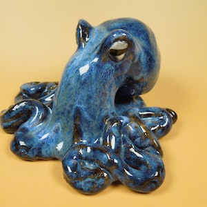 May include: A ceramic octopus figurine featuring a glossy, mottled glaze in shades of deep blue, navy, and hints of dark brown. The sculpture depicts the creature with a rounded head and multiple curled tentacles resting on a flat surface.