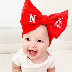May include: A baby wearing a white onesie is sitting on a white blanket. The baby is wearing a red bow headband with the letter "N" and the words "Go Big Red" embroidered on it.