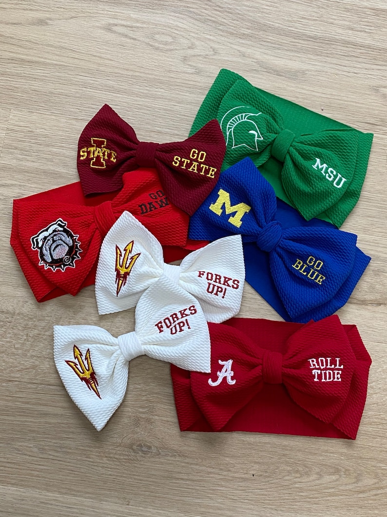 May include: A collection of colourful hair bows with various college logos and slogans, including "Go State", "MSU", "Go Dawg", "M", "Go Blue", "Forks Up!" and "Roll Tide".