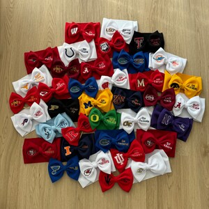 May include: A collection of 25 white, red and blue headbands with embroidered logos of various college and professional sports teams. The headbands are arranged in a semi-circle on a wooden surface.