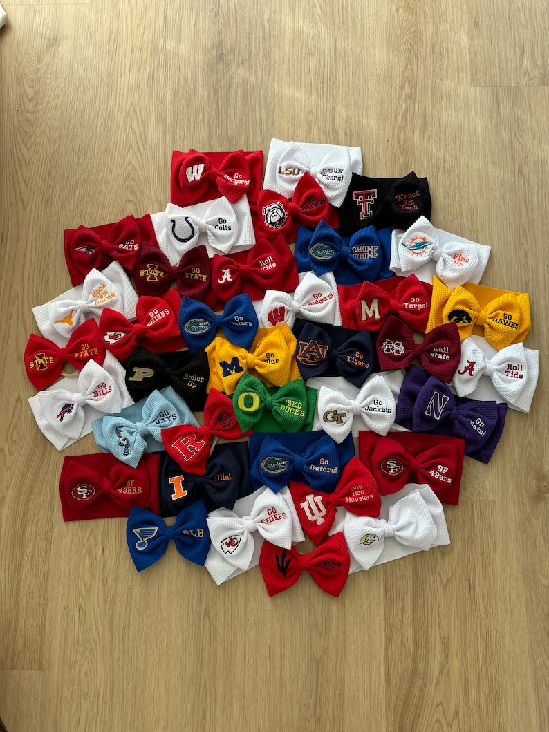 May include: A collection of 25 white, red and blue headbands with embroidered logos of various college and professional sports teams. The headbands are arranged in a semi-circle on a wooden surface.