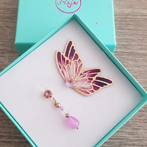 May include: A pair of earrings in a teal box. The earrings are a purple butterfly charm and a dangling purple stone with gold accents. The box has the word "Refe" written on it.