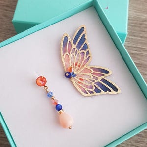 May include: A pair of earrings in a turquoise box. One earring features a butterfly design with pink, orange, and blue colors. The other earring is a teardrop-shaped pendant with a coral-colored stone and blue and clear crystals.