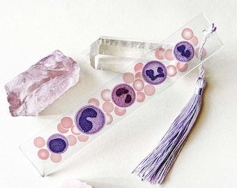 Puede incluir: Un marcapáginas transparente con un diseño de células de color morado y rosa. El marcapáginas tiene un borla morado.