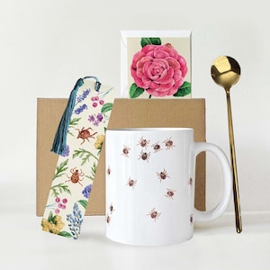 May include: A white ceramic mug adorned with brown insect illustrations, a gold spoon, a floral bookmark, and a greeting card featuring a pink rose. The items are arranged on a white surface.