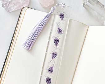May include: A clear acrylic bookmark with a lavender tassel. The bookmark features a series of purple, stylized parasite illustrations. The bookmark is resting on an open notebook, with crystals in the background.