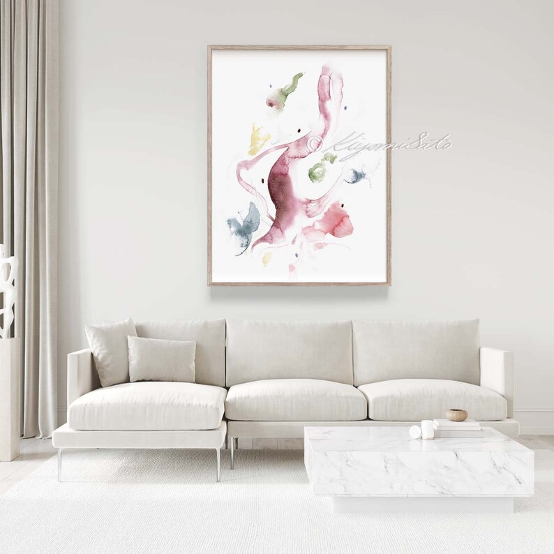 May include: Abstract watercolor painting in shades of pink, green, and yellow. The painting features a large, organic shape with smaller shapes scattered around it. The painting is framed in a light wood frame and is hanging on a white wall.
