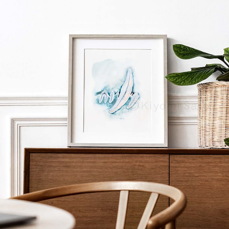 May include: A framed watercolor painting of abstract shapes in shades of blue and white. The painting is set against a white wall with a wooden cabinet and a potted plant in the background.