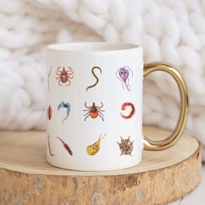 May include: White ceramic mug with a gold handle, decorated with colorful illustrations of various parasites. The mug is set on a wooden surface against a soft, white background. A unique and educational gift for science enthusiasts.
