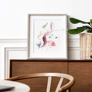 May include: A framed watercolor painting with abstract shapes in shades of pink, green, and blue. The painting is set against a white wall with a wooden cabinet and a potted plant in the background.