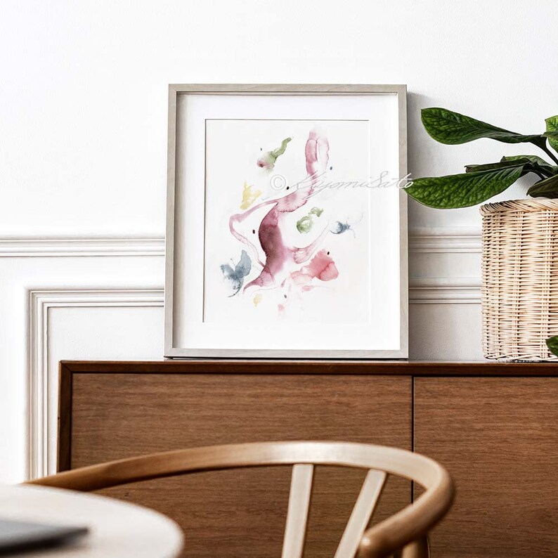 May include: A framed watercolor painting with abstract shapes in shades of pink, green, and blue. The painting is set against a white wall with a wooden cabinet and a potted plant in the background.