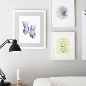 Op de afbeelding: Drie ingelijste prints op een witte muur. De print links toont een aquarel van twee kattenpootjes. De print in het midden toont een aquarel van een cirkelvormig patroon in tinten blauw, groen en paars. De print rechts toont een aquarel van een gele achtergrond met zwarte cijfers in een kolom.