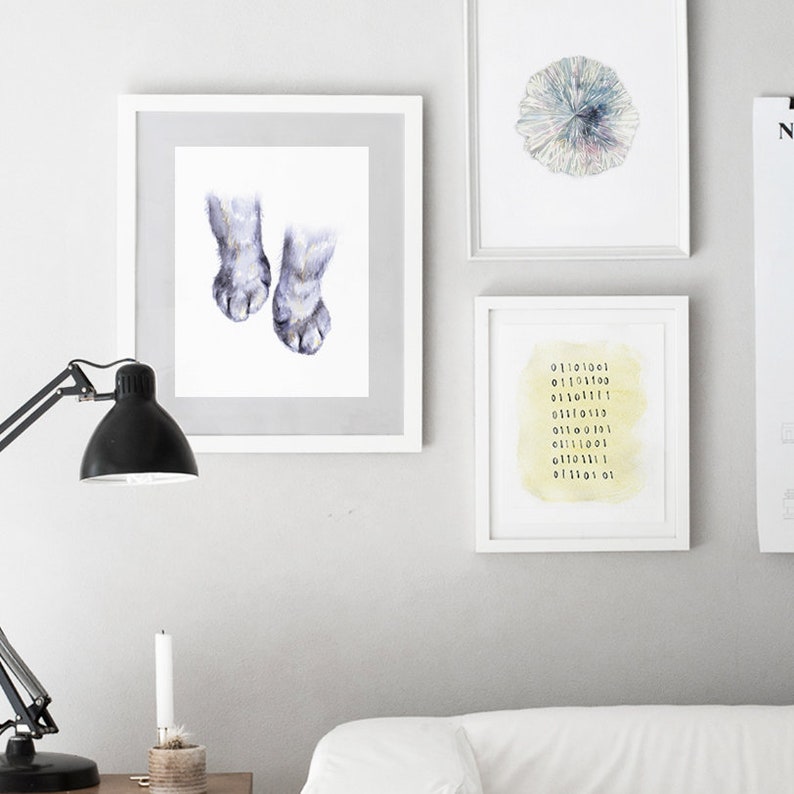 Op de afbeelding: Drie ingelijste prints op een witte muur. De print links toont een aquarel van twee kattenpootjes. De print in het midden toont een aquarel van een cirkelvormig patroon in tinten blauw, groen en paars. De print rechts toont een aquarel van een gele achtergrond met zwarte cijfers in een kolom.