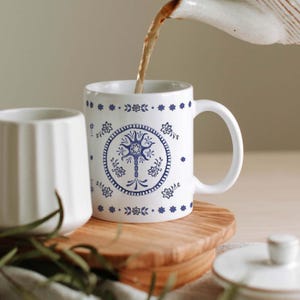 May include: A white ceramic mug featuring a blue circular pattern with a central tree-like design and small floral motifs. The mug has a handle on the right side and is being filled with a dark brown liquid poured from a white ceramic teapot above.