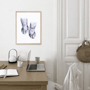 Op de afbeelding: Een aquarel schilderij van twee kattenpootjes met lichtgrijs haar en donkergrijze klauwen. Het schilderij is ingelijst in een licht houten lijst en hangt aan een witte muur.