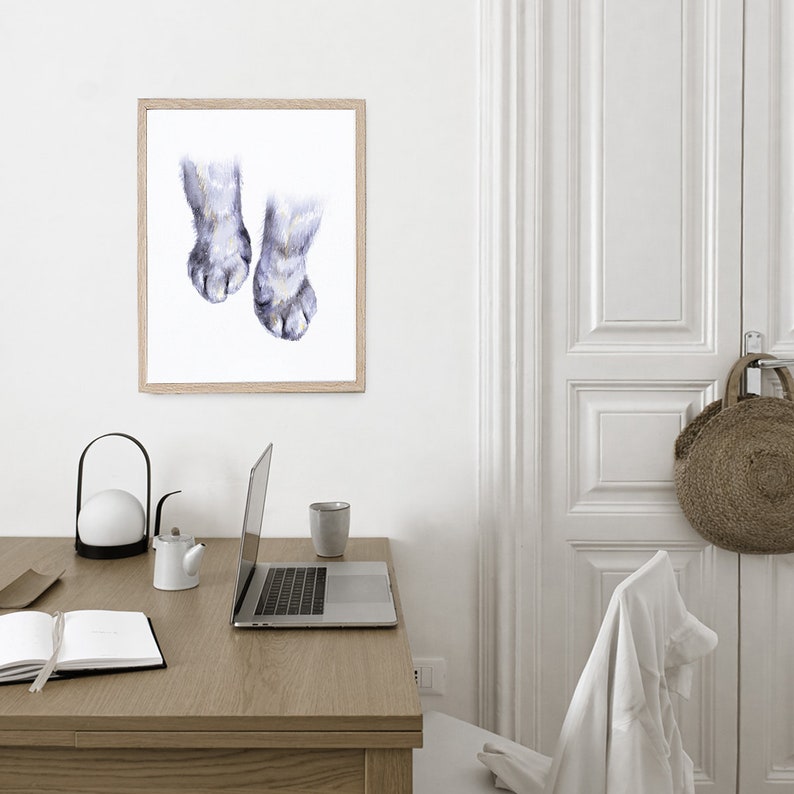 Op de afbeelding: Een aquarel schilderij van twee kattenpootjes met lichtgrijs haar en donkergrijze klauwen. Het schilderij is ingelijst in een licht houten lijst en hangt aan een witte muur.