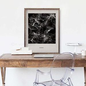 May include: A framed art print featuring a black and white abstract design resembling neural networks. The print is labeled "NEURON" and is displayed on a wooden desk with a laptop, a white box, and a clear plastic chair.