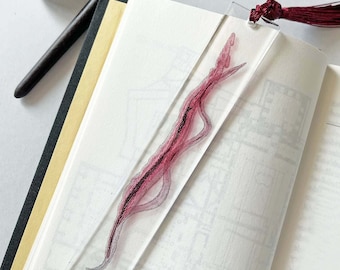 May include: A clear bookmark with a long, thin, pink and black object encased in resin. The bookmark has a red tassel.