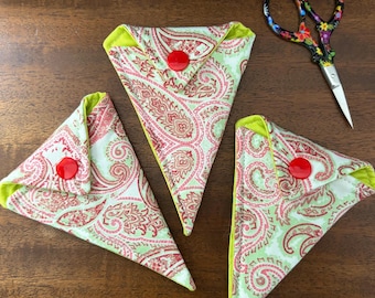Op de afbeelding: Drie scharen etuis met paisley print, rode knopen en limoengroene randen worden gepresenteerd op een houten oppervlak. Een decoratieve schaar met een bloemenmotief is ook aanwezig. De etuis zijn driehoekig van vorm.