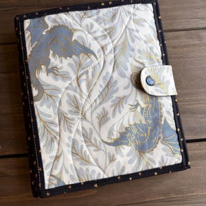 May include: A rectangular fabric book cover featuring a cream background with a repeating pattern of grey dragons and gold leaf-like foliage. The cover has black binding with small gold stars and a snap closure tab with a single dark button.