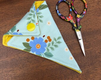 Op de afbeelding: Een kleine schaar met bloemenpatroon naast een lichtblauwe schaarkoker. De koker heeft een gele binnenkant en is versierd met blauwe bloemen en oranje accenten. De schaar heeft een kleurrijk bloemenpatroon op de handvatten.
