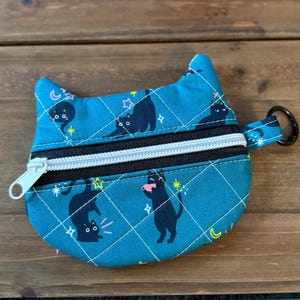 May include: A teal quilted coin purse shaped like a cat head with pointed ears. The fabric features a pattern of black cats, yellow stars, and pink stars. It includes a black zipper with a white pull and a black metal ring on the side.