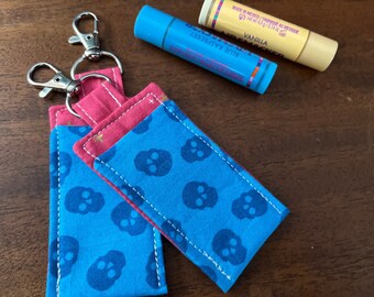 May include: Two keychains with a blue fabric with a skull pattern and a red fabric with a gold glitter pattern. The keychains have silver key rings.
