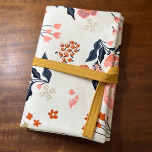 May include: Folded fabric item with a floral pattern in pink, orange, and navy blue on a cream background. A mustard yellow strap wraps around the item. The floral design includes flowers, leaves, and geometric shapes.