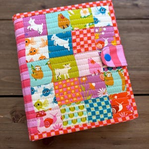 Könnte beinhalten: Eine farbenfrohe, gesteppte Notizbuchhülle mit Patchwork-Design. Die Hülle zeigt Quadrate mit Tier- und Blumenmustern in verschiedenen Farben, darunter Rosa, Blau, Grün und Orange. Sie hat einen Knopfverschluss und einen rot-weiß karierten Rand.
