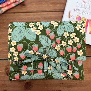 Op de afbeelding: Een gewatteerde boekomslag met een groene achtergrond, versierd met een aardbeien- en bloemenpatroon. De omslag is vastgebonden met een bijpassende strik. Extra stoffen met bloempatronen zijn zichtbaar.