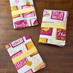 Puede incluir: Tres carteras de té de tela con fondo blanco, con diseños rosas, amarillos y morados. Cada cartera está envuelta con una banda elástica amarilla. Las carteras tienen texto que dice "Chamomile", "Oolong" y "Lemon".